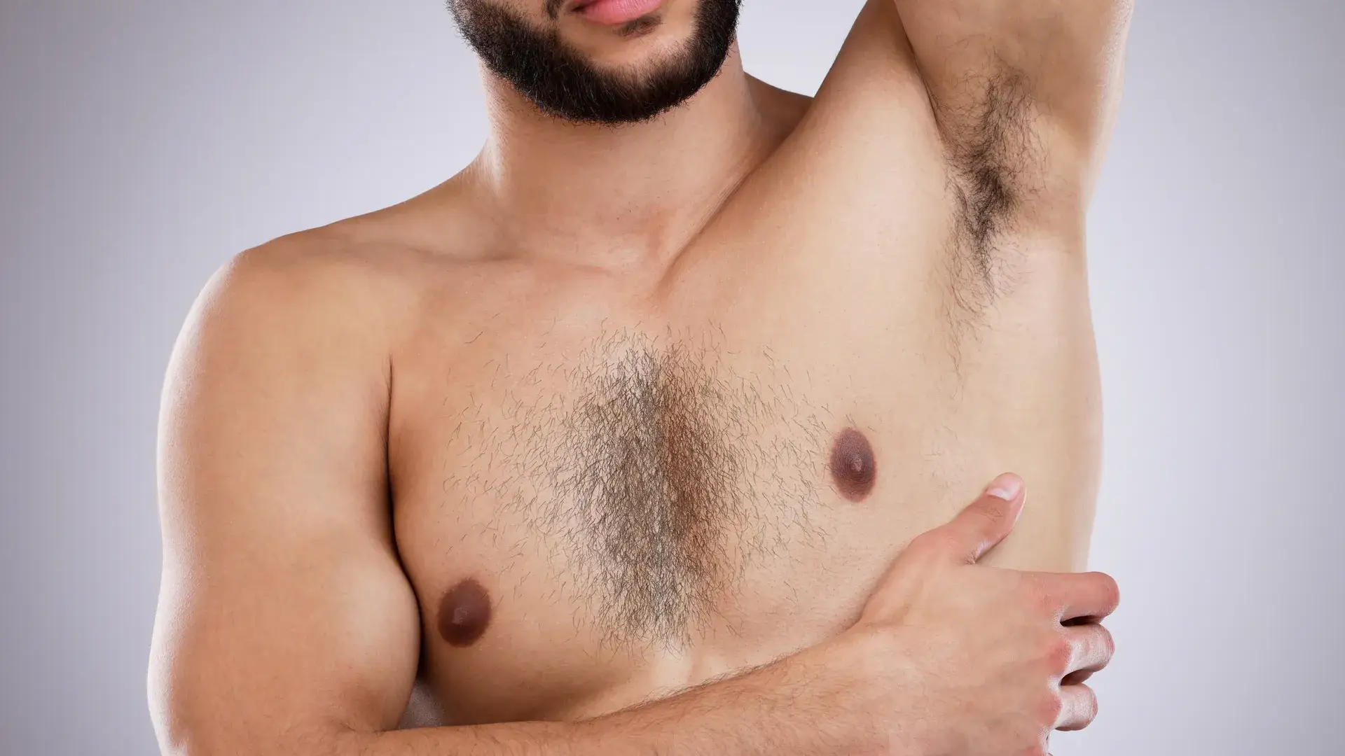 A shirtless man with a beard, one arm raised, showing underarm, chest and forearm hair against a grey background