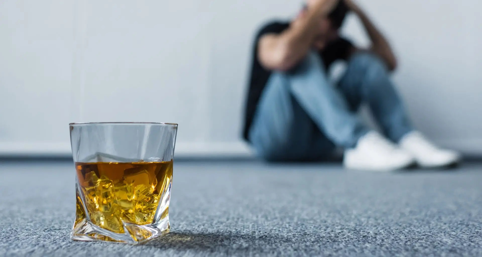 Close-up of a whiskey glass on the floor with a blurred background of a distressed man, underscoring the importance of alcohol testing in addressing substance abuse.