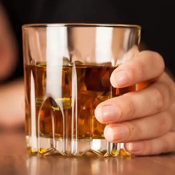 Close-up of a hand holding a whiskey glass with a blurred figure in the background, illustrating the personal impact of alcohol use, relevant in hair alcohol testing