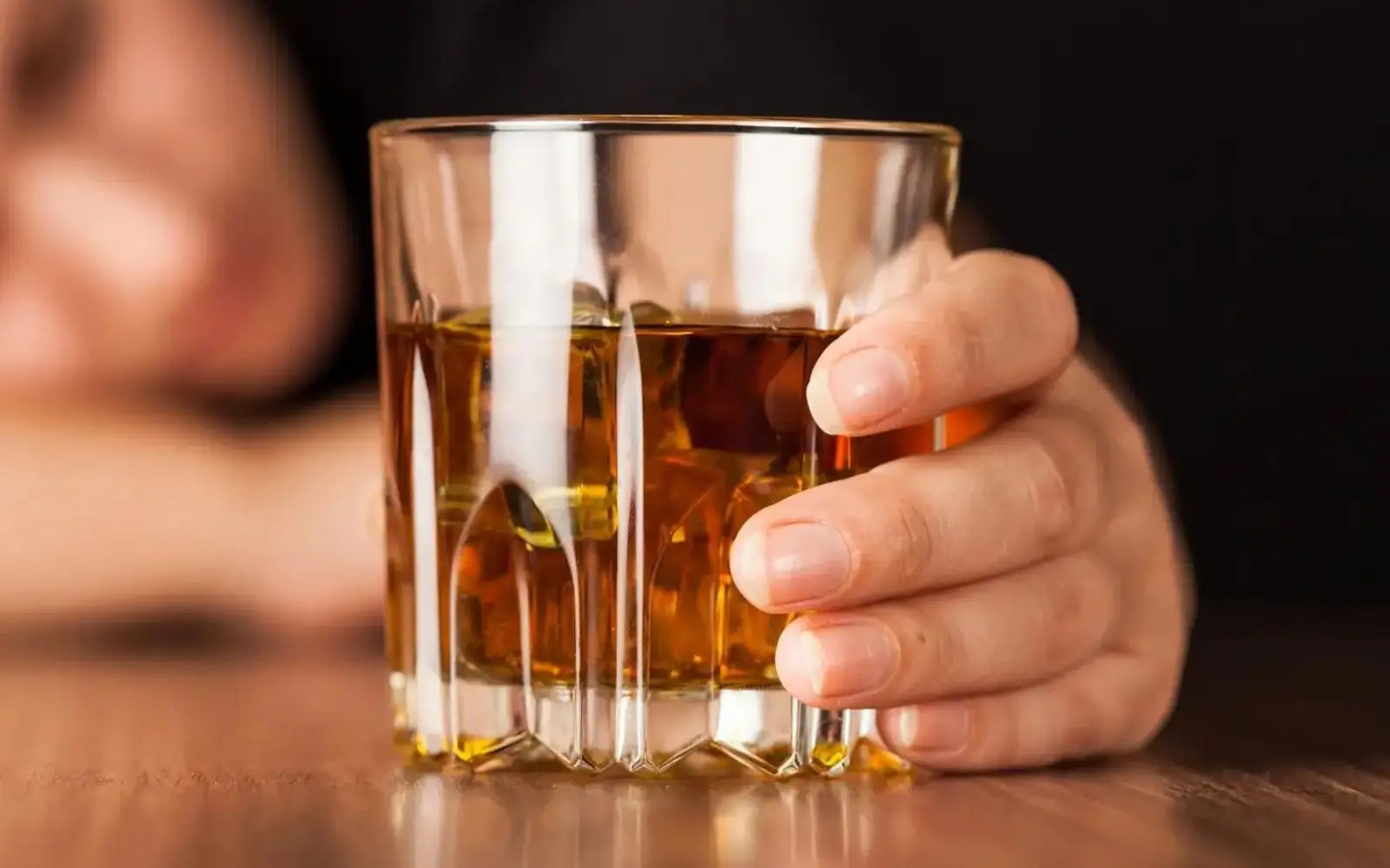 Close-up of a hand holding a whiskey glass with a blurred figure in the background, illustrating the personal impact of alcohol use, relevant in hair alcohol testing
