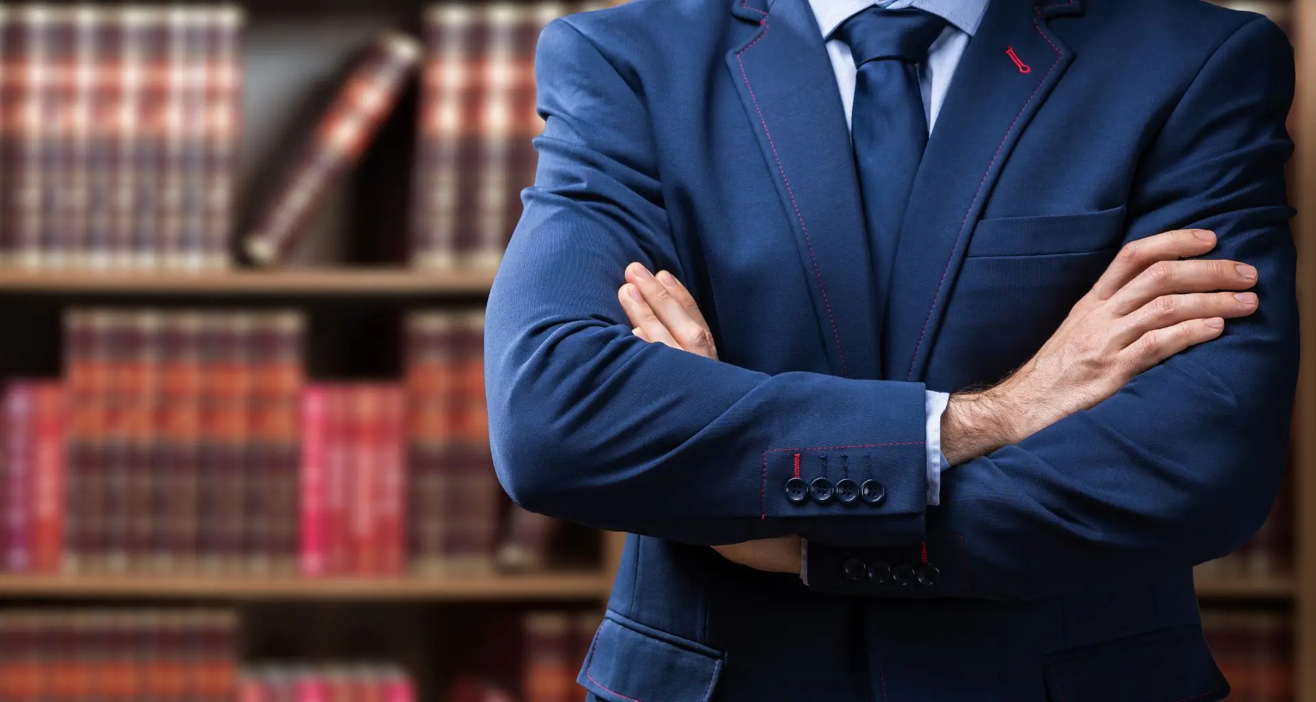 Confident legal expert in a blue suit standing with crossed arms in a library full of law books, representing professionalism in medico-legal consulting related to drug reports
