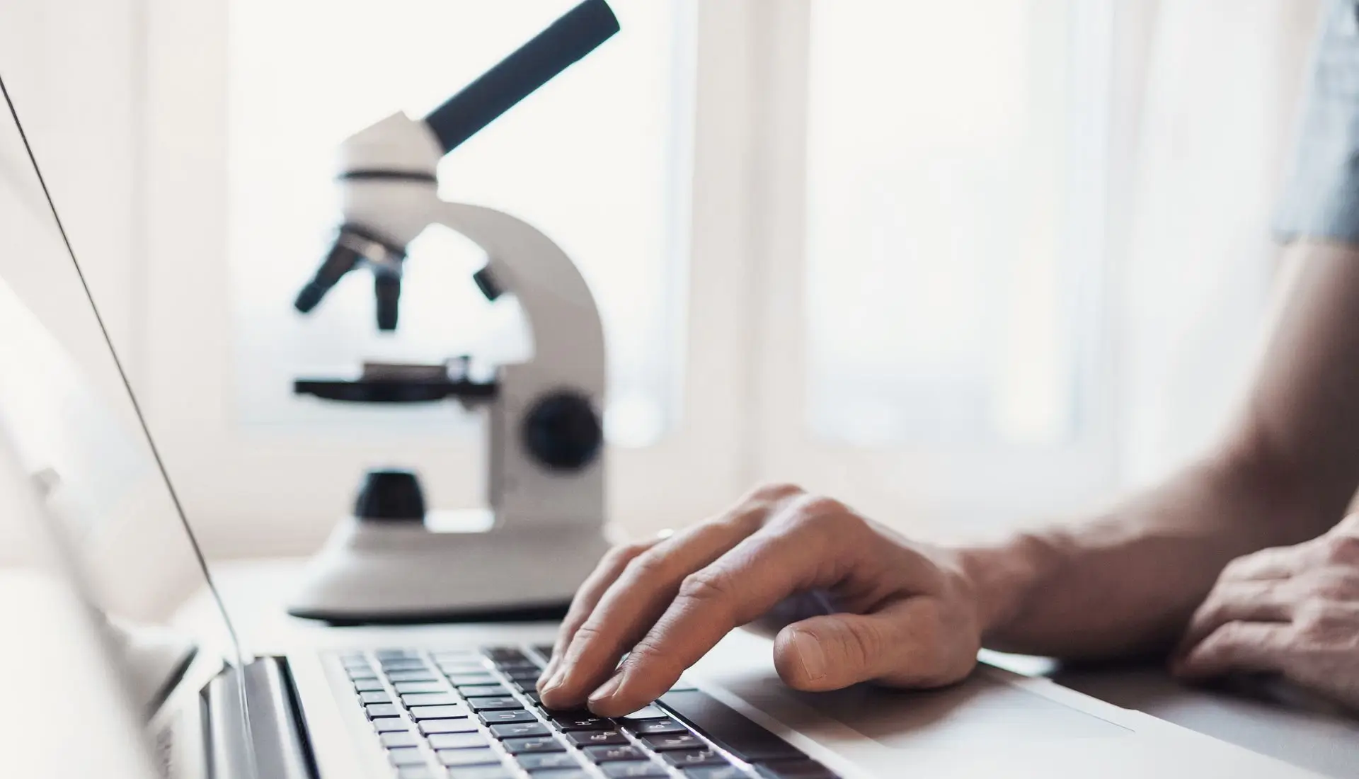 Forensic toxicologist using a laptop with a microscope in the background, symbolizing the integration of digital and traditional analysis in hair drug testing