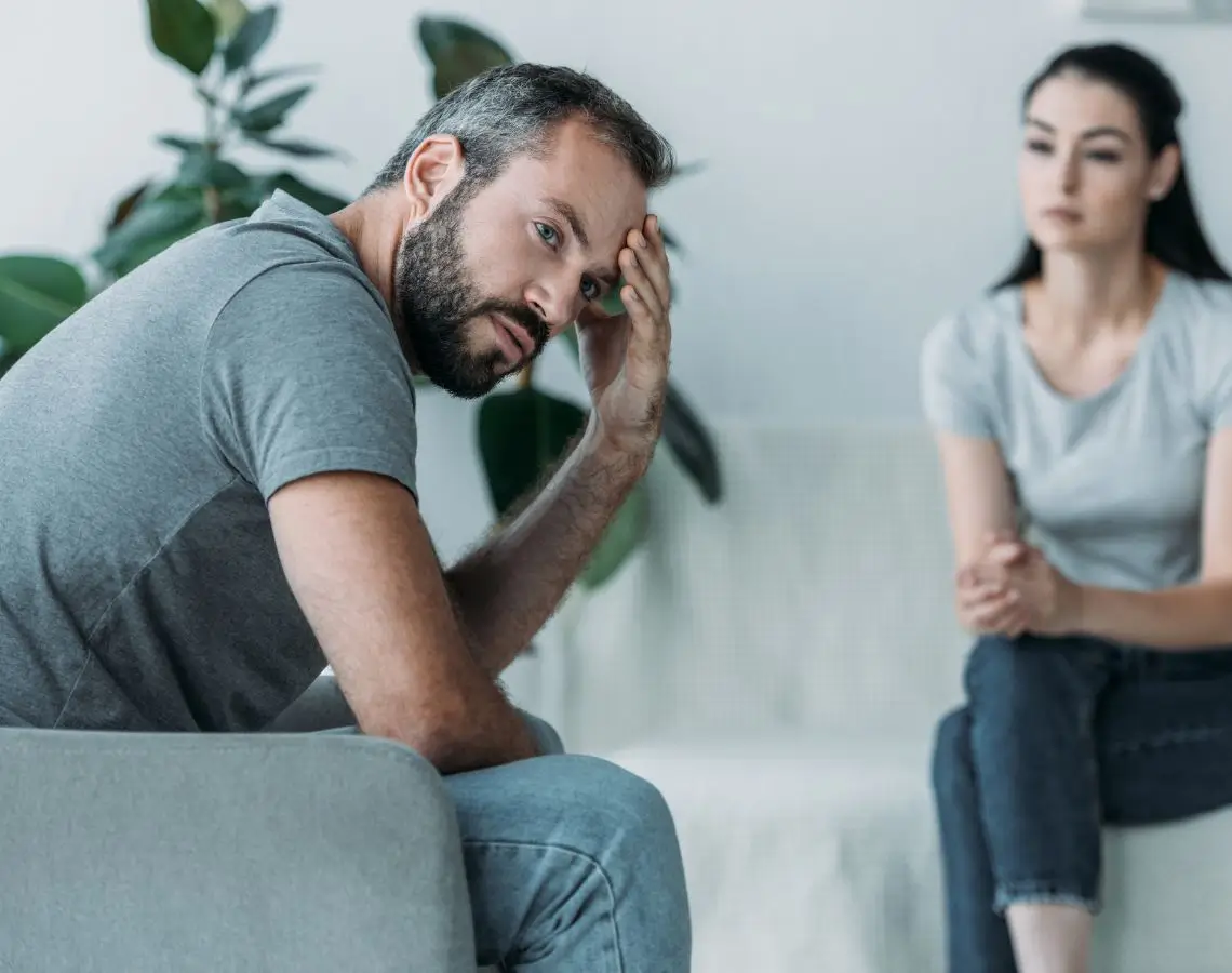 Distressed man sitting on a couch with a concerned woman in the background, illustrating the personal impact of addiction and the potential need for hair drug testing in family situations