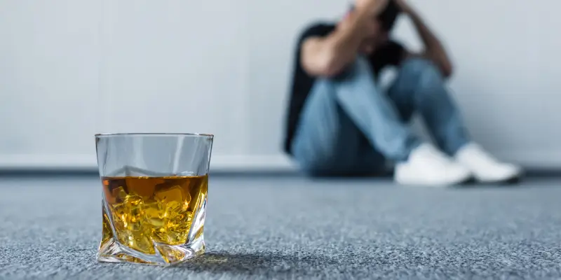 Close-up of a whiskey glass on the floor with a blurred background of a distressed man, underscoring the importance of alcohol testing in addressing substance abuse.