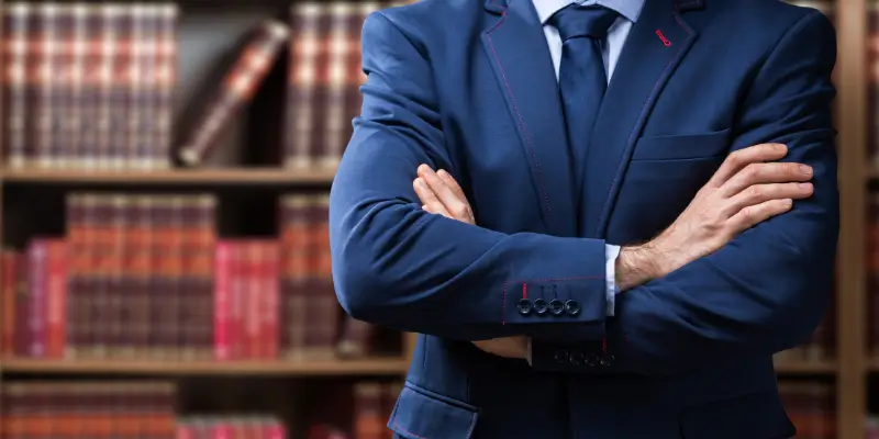 Confident legal expert in a blue suit standing with crossed arms in a library full of law books, representing professionalism in medico-legal consulting related to drug reports