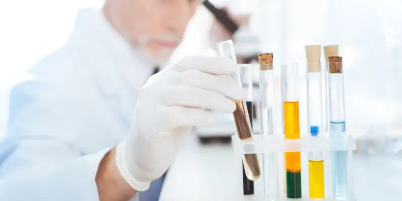 Blurry background image of a scientist in a lab coat analyzing a hair sample among test tubes with colourful solutions, focusing on precision in drug testing.